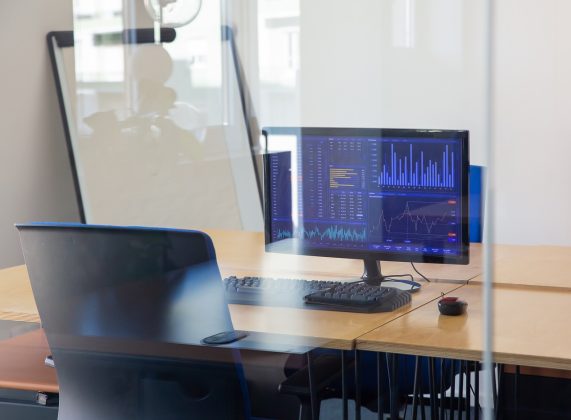 Empty traders workplace behind glass wall. Office room with flipchart, desk with chair and computer. Trading charts on monitor. Stock market exchange concept